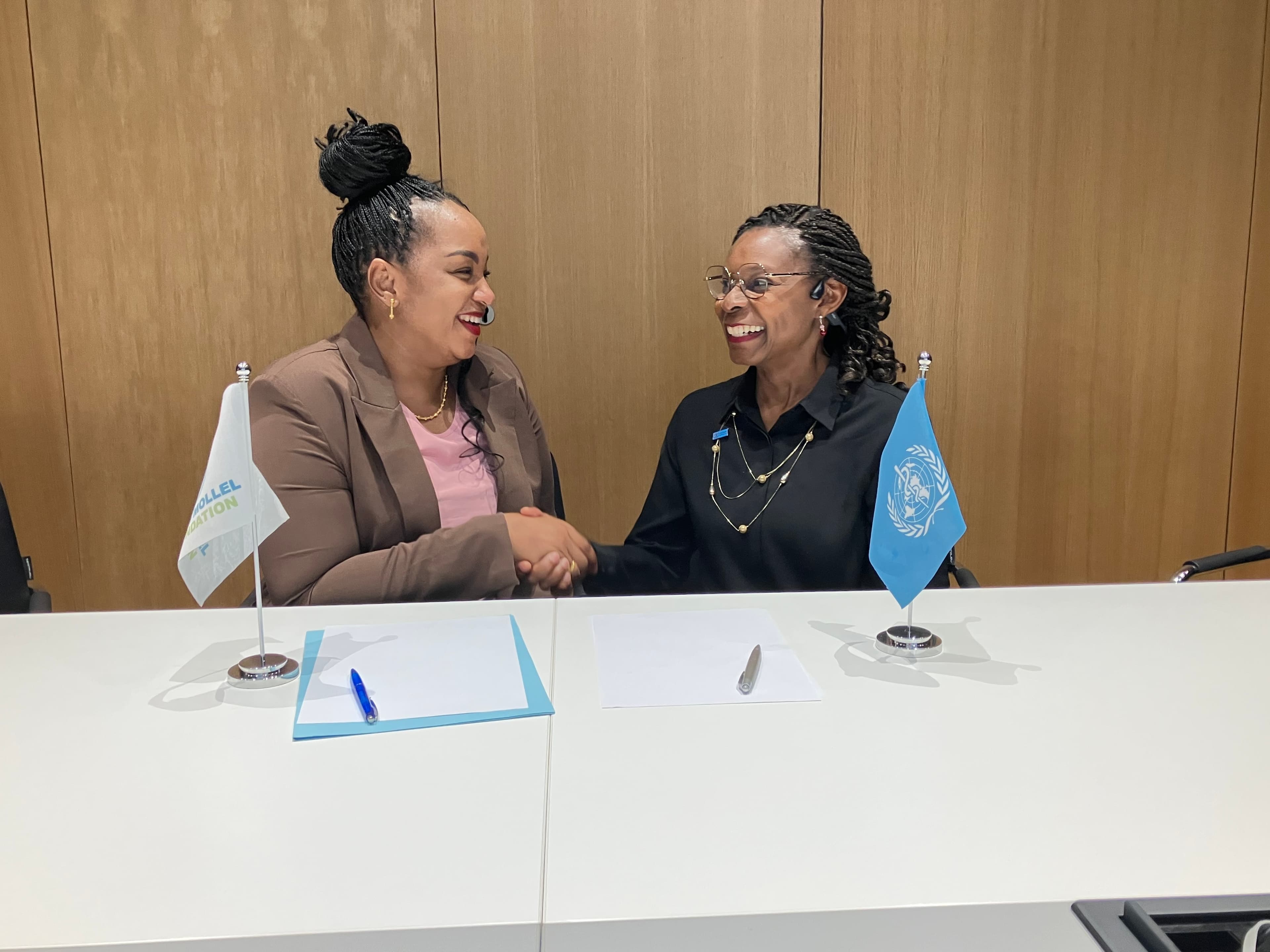 Doris Mollel shakes hands with a WHO representative across the signing table, with the foundation and WHO flags
